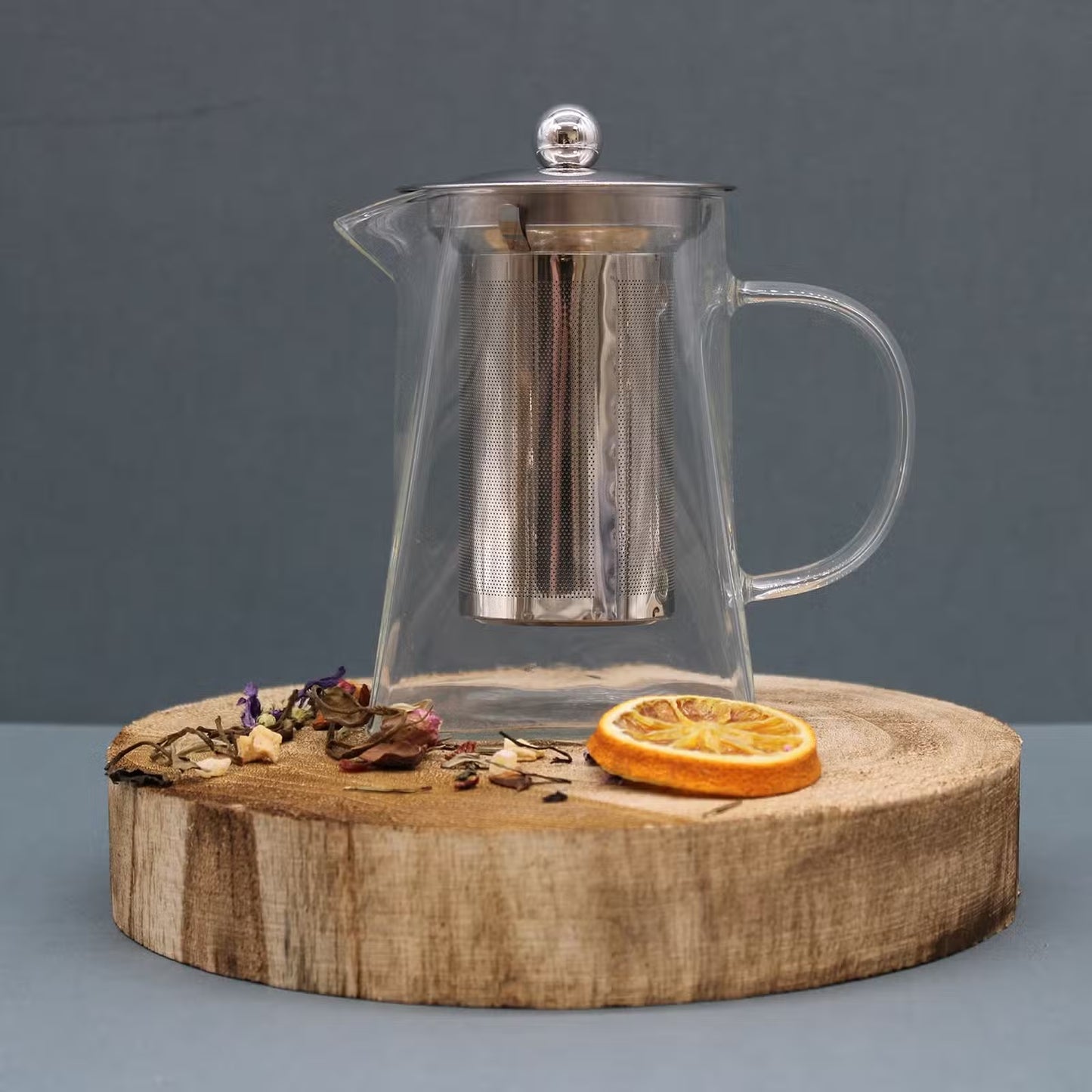 Clear glass teapot with metal mesh on a wooden stand against a gray background - Kraas