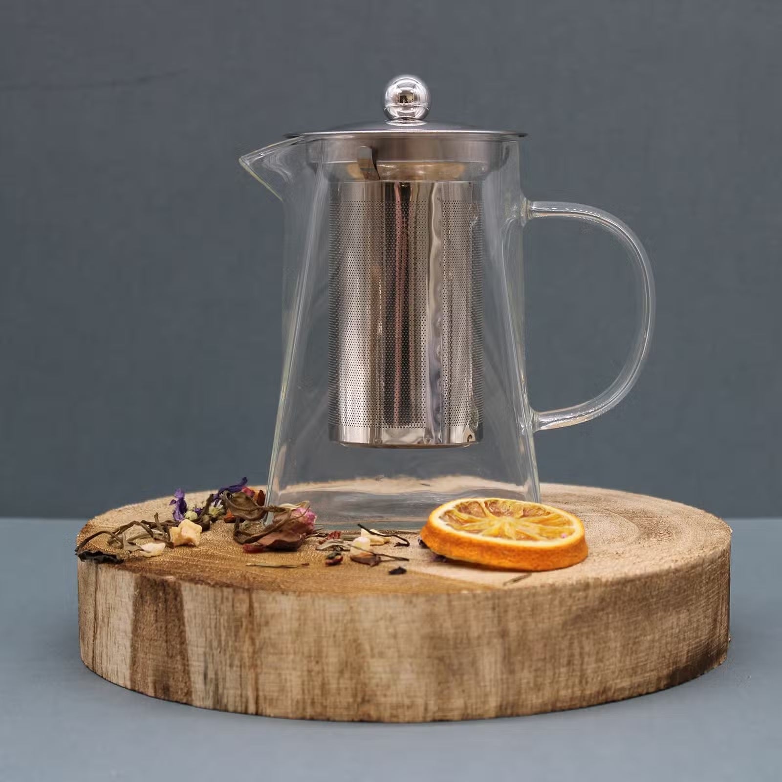 Clear glass teapot with metal mesh on a wooden stand against a gray background - Kraas